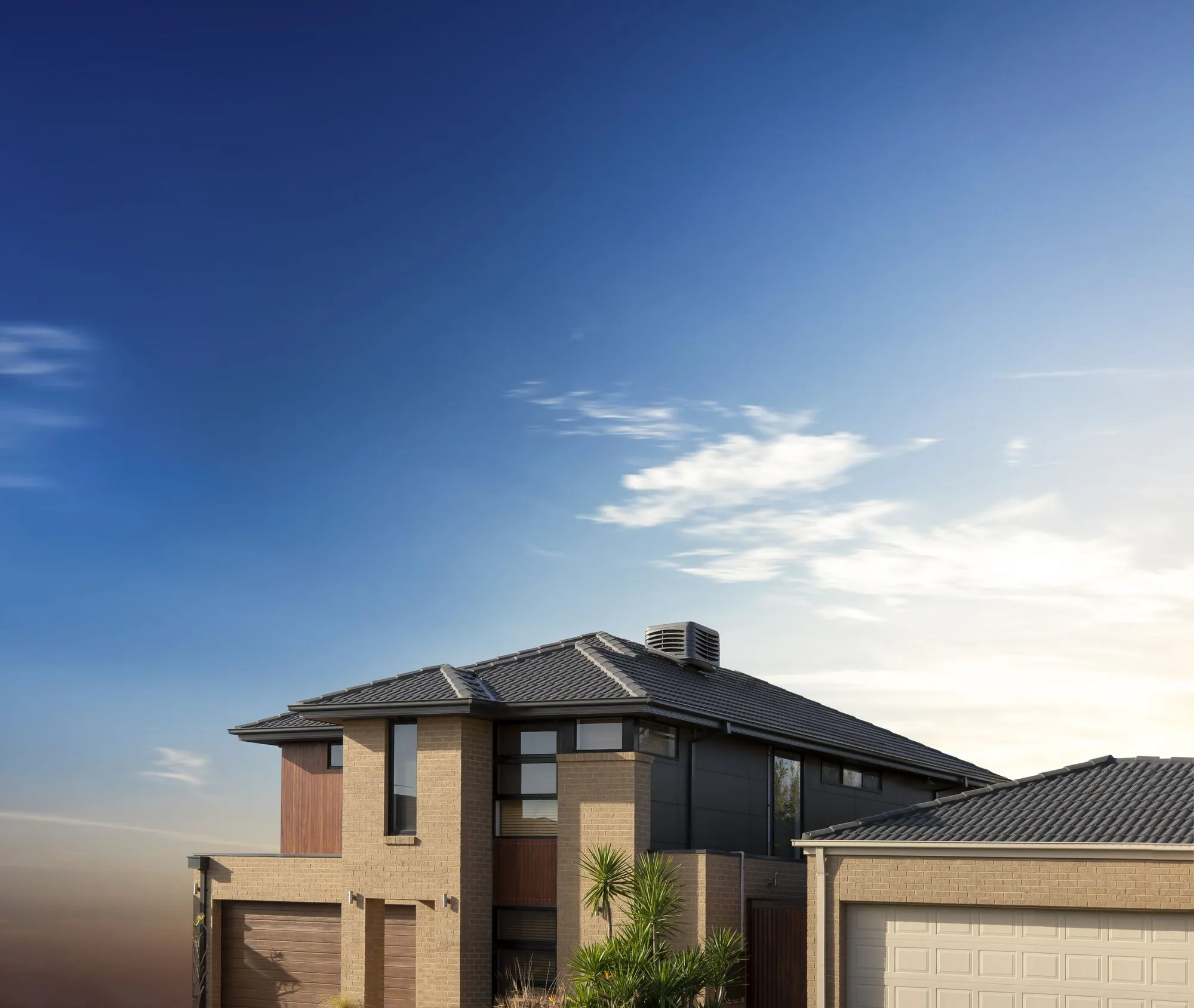 Brivis Contour evaporative cooler in charcoal installed on the tiled roof of a brick home under a blue sky